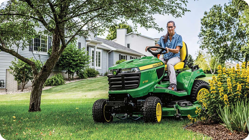 Woman in the seat of a John Deere riding lawn mower cutting grass.